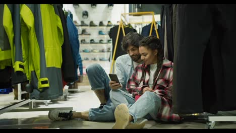 Couple-in-the-sport-department-store-sitting-on-the-floor,-watching-mobile-phone