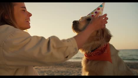Shooting-close-up:-a-blonde-girl-wishes-her-dog-a-happy-birthday-on-a-sunny-beach-and-puts-a-festive-cap-on-her