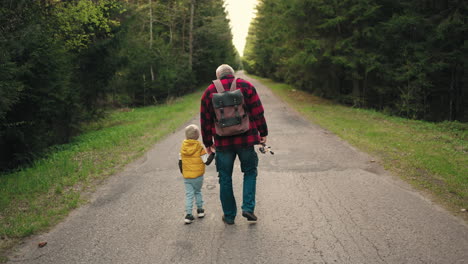old-man-with-backpack-is-holding-hand-of-his-little-grandson-walking-together-on-road-across-forest-rear-view