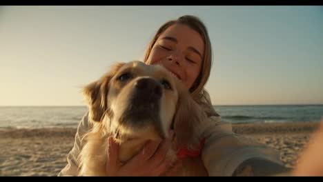 View-from-the-side-of-the-phone,-a-blonde-girl-and-her-light-dog-take-a-selfie-on-the-background-of-a-sunny-beach-in-the-morning,-close-up-shot