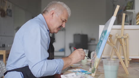 An-elderly-man-in-an-apron-paints-a-picture-with-a-brush-while-sitting-at-the-table
