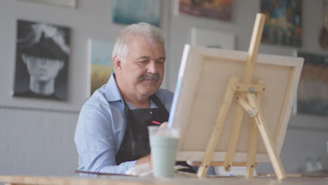 An-elderly-man-in-an-apron-paints-a-picture-with-a-brush-while-sitting-at-the-table