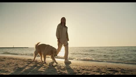 A-blonde-girl-walks-along-with-a-light-coloring-big-dog-on-a-sunny-beach-in-the-morning