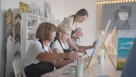 High-angle-view-of-cheerful-senior-friends-painting-on-canvas.-Senior-woman-smiling-while-drawing-with-the-group.-Seniors-Attending-Painting-Class-Together.-Senior-men-having-fun-painting-in-art-class