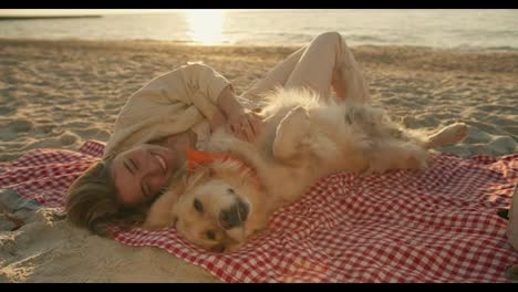 Close-up-shot:-a-blonde-girl-with-her-light-colored-dog-lie-on-a-mat-on-a-sunny-beach-in-the-morning.-The-girl-strokes-the-dog-and-she-is-happy