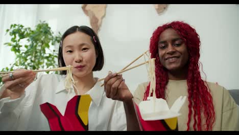Diverse-female-friends-eating-noodles-together-at-home-using-chopsticks,-positive-mood