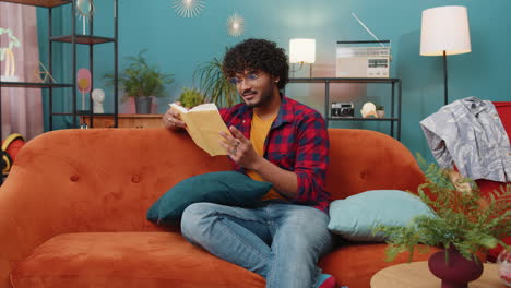 Indian-man-reading-interesting-book-turning-pages-smiling-enjoying-literature-taking-a-rest-on-sofa