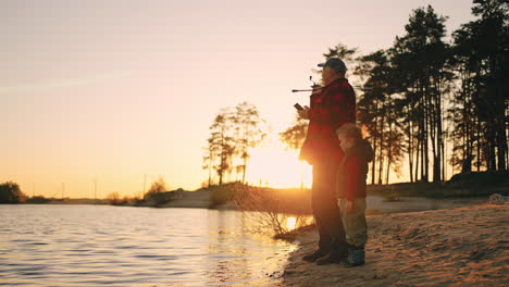 happy-grandfather-and-grandson-are-fishing-on-river-shore-in-picturesque-natural-landscape-with-pines