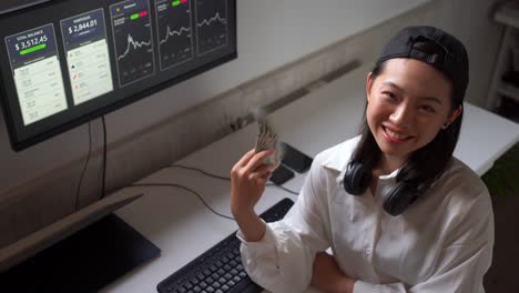 Smiling-Asian-trader-cooling-down-with-paper-dollars-against-monitor