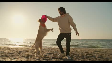 Der-Typ-Spielt-Morgens-Mit-Seinem-Hellen-Hund-An-Einem-Sonnigen-Strand-Mit-Einem-Roten-Spielzeug