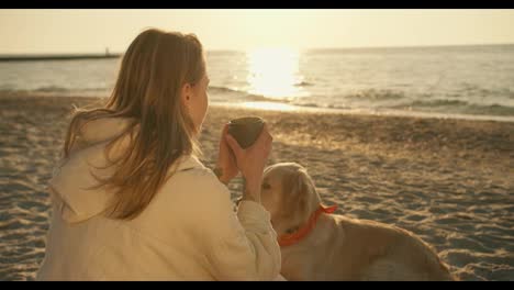 A-blonde-girl-drinks-hot-tea-from-a-mug-of-a-thermos-on-a-sunny-beach-in-the-morning.-Near-the-girl-is-her-big-cute-dog-of-light-coloring