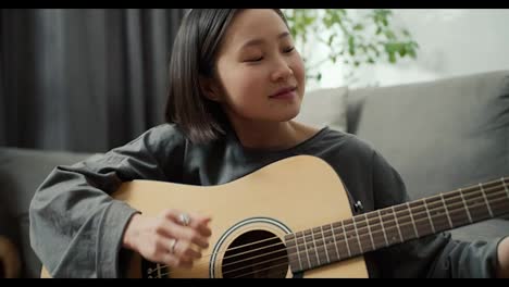 Chica-Asiática-Tocando-Instrumento-Musical-De-Guitarra-Acústica-En-Casa,-Sentada-En-El-Suelo,-De-Cerca