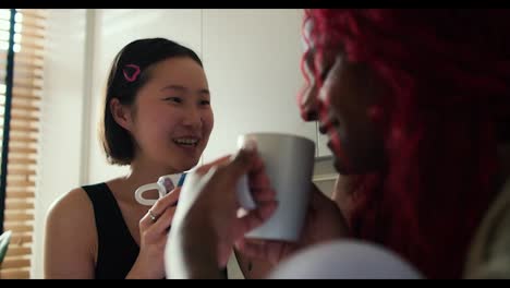 Two-beautiful-women-multiethnic-friends-drinking-coffee-while-talking-in-the-kitchen-at-home,-close-up