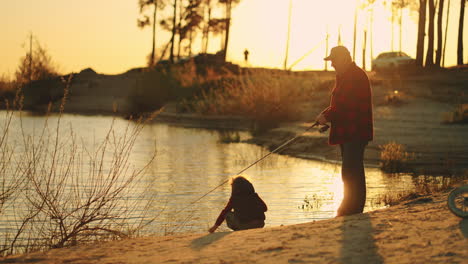 old-fisherman-and-little-boy-are-spending-time-on-shore-of-lake-or-river-family-fishing-in-sunset