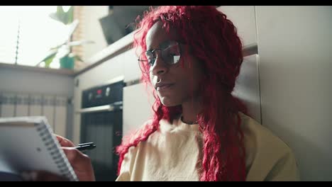 Afroamerikanisches-Mädchen-Mit-Roten-Lockigen-Haaren,-Das-Auf-Dem-Küchenboden-Studiert-Und-Notizen-Macht
