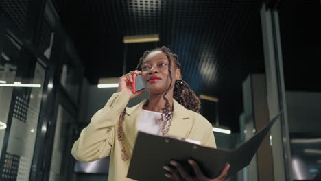 African-Business-woman-talking-smartphone-in-office-interior.-Smiling-black-businesswoman-calling-mobile-phone-in-corridor.-Portrait-of-friendly-african-american-walking-in-business-center-hallway.