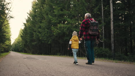 old-fisherman-and-his-little-grandson-are-walking-on-road-across-forest-in-morning-granddad-is-carrying-rod