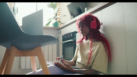 Afroamerikanisches-Mädchen-Mit-Roten-Locken,-Das-Mit-Einem-Laptop-Auf-Dem-Küchenboden-Sitzt-Und-Notizen-Macht