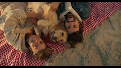 Top-view:-Happy-couple-on-the-beach-smiling-and-looking-at-the-camera.-Guy-and-girl-with-their-dog-posing-and-looking-at-the-camera-while-relaxing-on-a-sunny-beach-in-the-morning