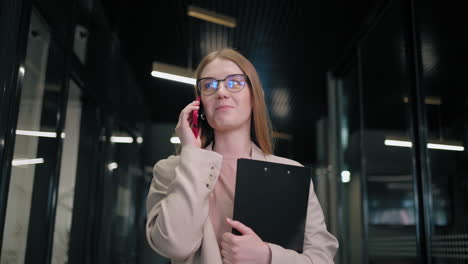 Portrait-of-a-business-woman-with-glasses-walking-in-the-office-corridor-and-talking-on-the-phone-with-clients