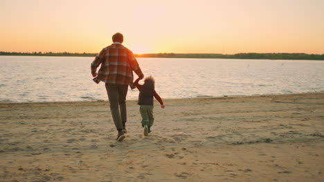 Ein-Glücklicher-Kleiner-Junge-Und-Sein-Vater-Rennen-Im-Sonnenuntergang-über-Das-Flussufer-Und-Freuen-Sich-über-Ein-Familienwochenende