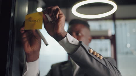African-American-businessman-working-with-sticky-notes-in-office.-Entrepreneur-explaining-business-plan.-Businessman-building-mind-map-with-sticky-notes-on-glass