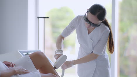 Beautician-doctor-doing-elos-hair-removal-depilation-on-attractive-young-woman-legs.-woman-legs-getting-pulses-of-laser-light-destroy-hair-follicle.-Laser-hair-removal-procedure-beautician-salon.