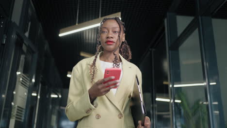Portrait-of-a-black-business-woman-walking-down-the-corridor-of-a-business-center-and-typing-a-message-in-her-phone