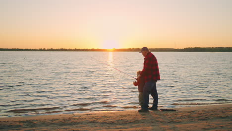 old-fisher-and-his-son-or-grandchild-are-catching-fish-by-fishing-rod-on-shore-of-lake-or-river