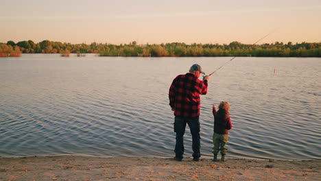 calm-fishing-on-river-shore-grandfather-and-little-grandson-are-catching-fish-by-rod-rear-view
