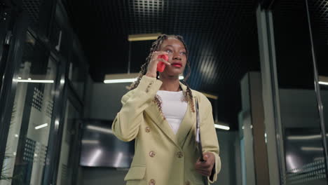 Portrait-of-a-black-business-woman-walking-down-the-corridor-of-a-business-center-talking-on-the-phone-and-carrying-documents