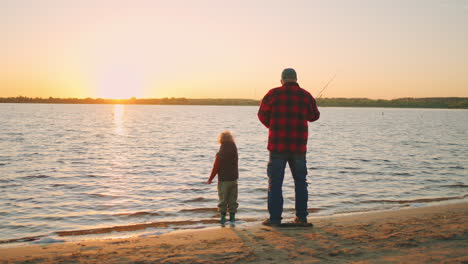 rest-on-river-shore-in-summer-sunset-old-fisher-and-his-grandson-are-fishing-in-evening-rear-view
