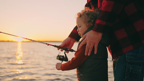 happy-curly-toddler-is-catching-fish-by-fishing-rod-father-or-grandpa-is-helping-teaching-son-to-fish