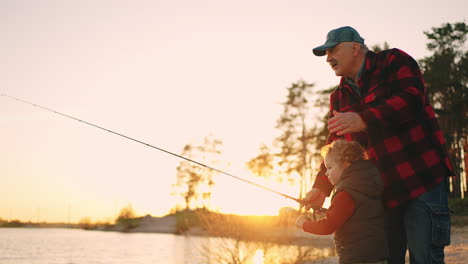 careful-grandfather-and-little-grandson-are-fishing-together-in-river-shore-in-sunset