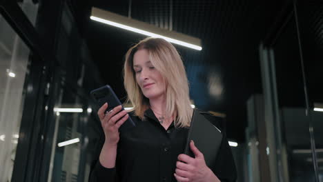 Smiling-businesswoman-looking-on-cellphone-indoors.-Surprised-business-woman-reading-message-on-mobile-phone-in-office-corridor.-woman-looking-smartphone-screen-in-business-center.-walking-office