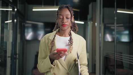 Portrait-of-a-black-business-woman-walking-and-flipping-through-the-news-feed-on-the-phone-in-the-office-corridor