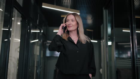 A-blonde-business-woman-of-30-40-years-old-in-a-black-shirt-walks-down-the-corridor-of-the-office-and-talks-on-the-phone