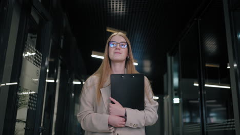A-business-woman-with-glasses-walks-down-the-corridor-of-a-business-center-with-papers