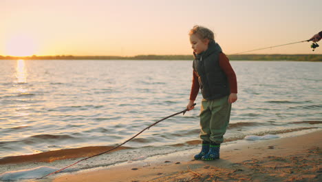 little-curly-boy-is-playing-with-stick-on-river-shore-touching-water-resting-with-his-father-fisher