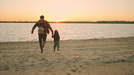 joyful-father-and-little-son-are-resting-and-having-fun-on-river-shore-running-to-water-in-sunset
