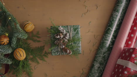 top-view-of-the-decorator's-desktop-Fully-visible-the-table-with-the-decorations.-Female-hands-put-and-finalize-Christmas-gift-wrapped-in-craftool-paper-on-a-wooden-table
