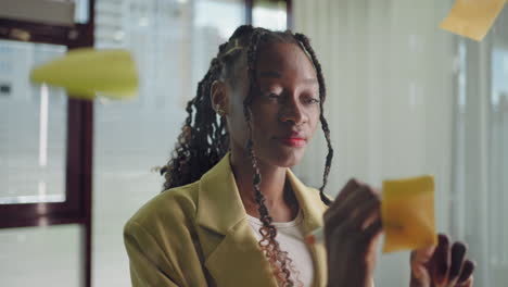 African-american-business-woman-using-sticky-notes-brainstorming-problem-solving-strategy-on-glass-whiteboard-leader-woman-showing-solution-for-project-deadline-in-office.