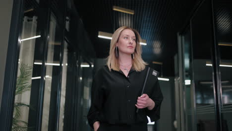A-happy-blonde-business-woman-of-30-40-years-in-a-black-shirt-walks-down-the-corridor-of-the-business-center-carrying-documents
