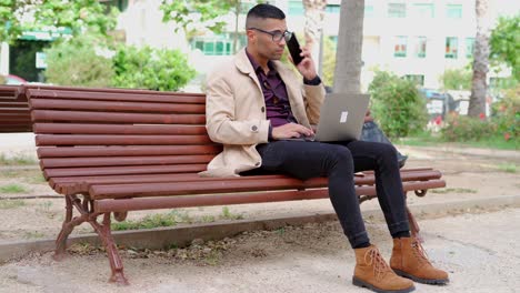 Young-businessman-in-trendy-outfit-speaking-on-phone-on-bench