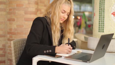 Mujer-Independiente-Escribiendo-Notas-Mientras-Trabaja-Con-Una-Computadora-Portátil-En-La-Cafetería.