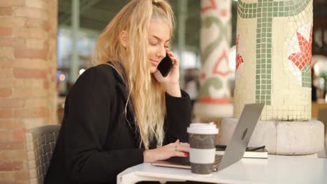 Mujer-Hablando-Por-Teléfono-Inteligente-Mientras-Trabaja-Con-Una-Computadora-Portátil-En-La-Cafetería