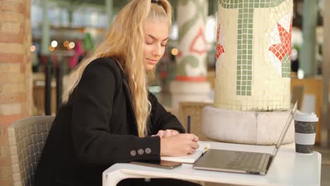 Mujer-Independiente-Escribiendo-Notas-Mientras-Trabaja-Con-Una-Computadora-Portátil-En-La-Cafetería.