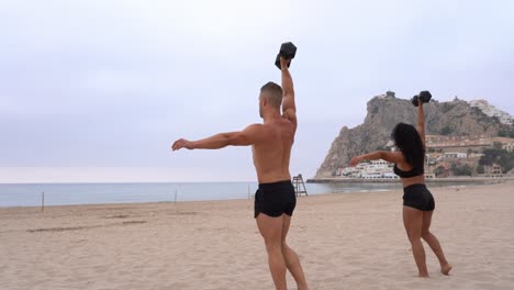 Diverse-strong-sportsman-and-sportswoman-doing-exercises-with-dumbbells-on-beach-at