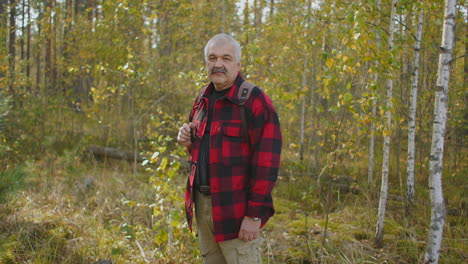 cheerful-middle-aged-man-is-standing-in-autumn-forest-with-yellowed-trees-at-daytime-smiling-to-camera-hiker-and-traveler