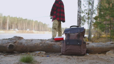 man-is-fishing-in-lake-standing-on-coast-in-forest-backpack-with-equipment-and-baits-on-groundrelax-hobby